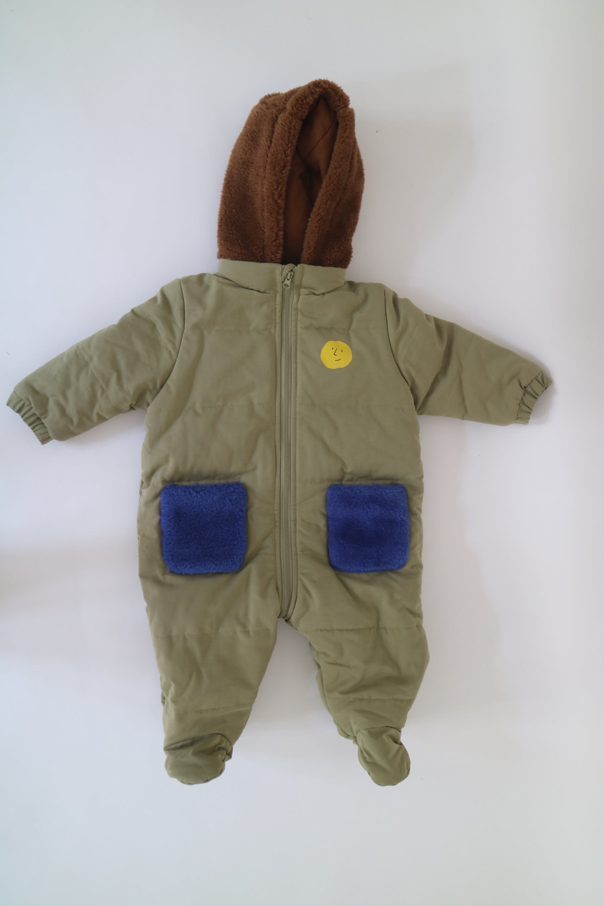 Fodrad overall i ljus militärgrön färg med kornblåa fickor och brun luva i teddy-material från Bobo Choses, modell Face Multicolor Overall. 